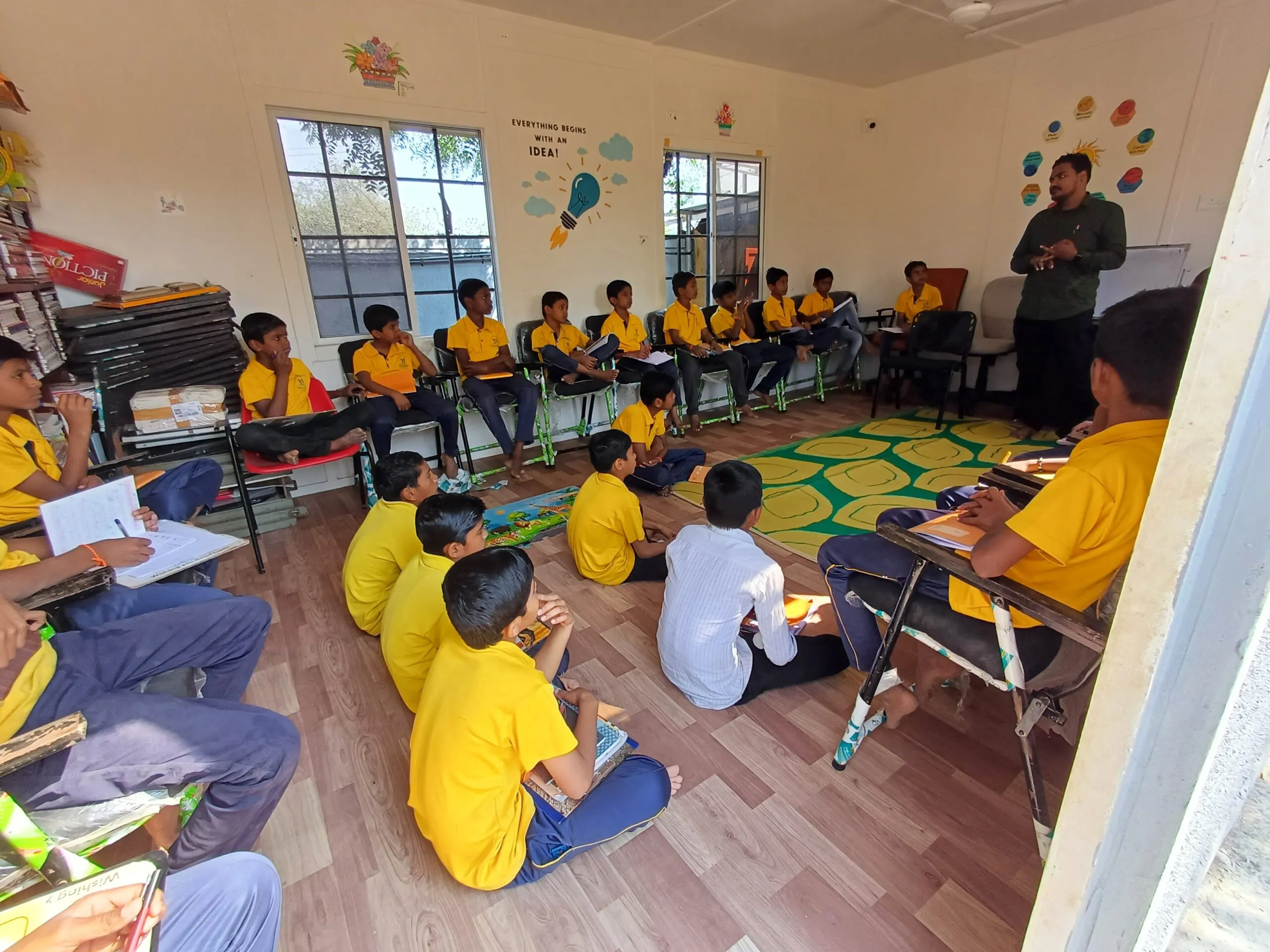 A classroom setting with a group of young students, mostly boys, dressed in yellow shirts sitting in a circular arrangement on chairs and the floor, attentively listening to a male teacher who is standing and speaking at the front. The classroom walls are decorated with educational posters, including one that reads "Everything begins with an IDEA!" featuring a lightbulb illustration. The room has a wooden floor and large windows allowing natural light to come in.