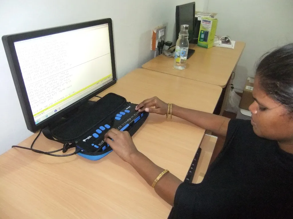 A visually impaired woman is using a refreshable braille display device connected to a desktop computer. She is typing on the braille keyboard while a monitor in front of her displays text in large font. The setup is on a wooden desk with other computers and items like a water bottle and electronics visible in the background. This image represents digital accessibility and inclusive technology for the visually impaired.
