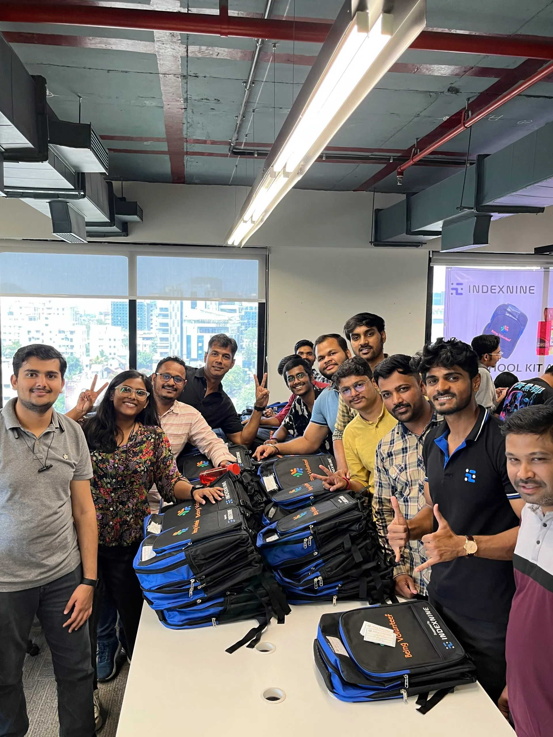 A group of enthusiastic professionals from Indexnine are gathered around a table inside a modern office space. The table is stacked with branded blue and black backpacks labeled "Project Yugankur." Several team members are flashing peace signs and smiles, clearly excited and proud. A banner in the background displays the Indexnine logo and mentions a "School Kit" initiative, suggesting a community outreach or CSR activity.