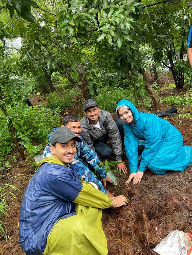 A cheerful group of four volunteers, wearing raincoats and smiling, are seen crouching in a lush green forest while planting a sapling. The scene suggests a tree plantation drive, with wet soil and rain-drenched foliage indicating it’s a monsoon day. Their hands are gently pressing the soil around the sapling, reflecting a shared commitment to environmental sustainability.