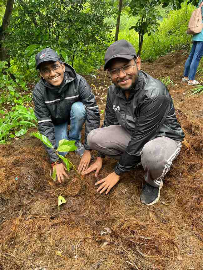 Two young men are kneeling in a forested area, smiling at the camera while planting a sapling together. They are dressed in black rain jackets and caps, with damp surroundings indicating recent rain. The green sapling they are planting is surrounded by brown mulch, emphasizing their involvement in a tree plantation or eco-volunteer initiative.