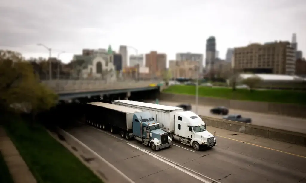 Wide urban cityscape showing semi-trucks driving on a highway beneath an overpass, with a downtown skyline, church, cars, and overcast sky in the background.